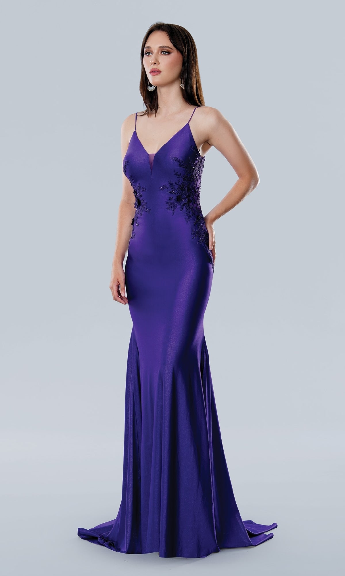 A woman stands in the Stella Couture 25186, a fitted floor-length purple prom dress with a v-neckline and floral lace on the bodice and sides, set against a plain light gray background.
